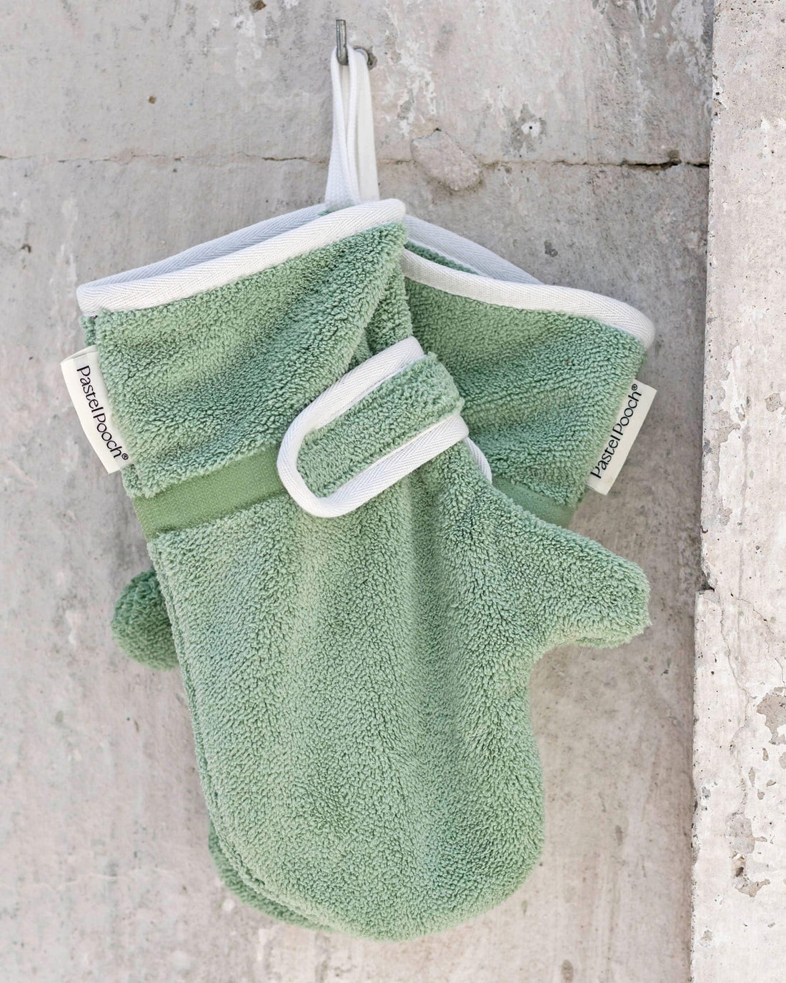 Pair of green bath mitts with white accents hanging on a concrete wall.